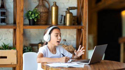 10 years old girl in white headphones during an online lesson. She does her homework online and...