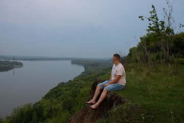 A young man sits on a cliff with bare feet in shorts and looks into the distance at a large river.