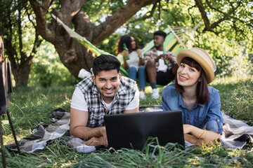 Smiling diverse couple using modern laptop while relaxing together on blanket at garden. Blur background of african couple sitting in hammock and chatting. Friends on nature.