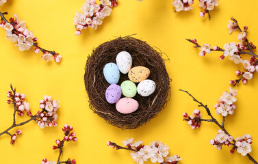 Obraz na płótnie Canvas Colored eggs in nest with beautiful pink flowering branches on yellow background. Springtime, easter concept. Flat lay, top view