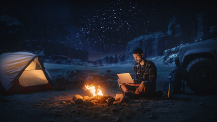 Night Tent Camping in Canyon: Male Traveler Uses Laptop Computer Sitting by Campfire. Man on Digital Remote Work, e-shopping, ecommerce, Using Internet, Social Media Posting on Vacation Trip  - obrazy, fototapety, plakaty