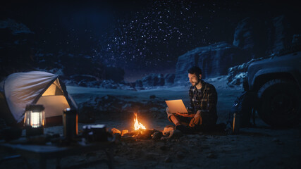 Male Traveler Sitting by Campfire Uses Laptop Computer while Tent Camping in the Canyon. Man doing...