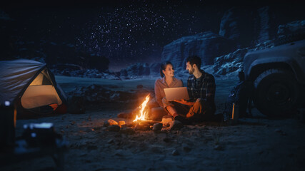 Happy Couple Camping in the Canyon at Night, Use Laptop Computer Sitting by Campfire. Two people Post Inspirational on Social Media, Use Internet, Watch Favourite Show on Streaming Service
