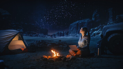 Night Tent Camping in Canyon: Female Traveler Uses Laptop Computer Sitting by Campfire. Woman on Digital Remote Work, e-shopping, ecommerce, Using Internet, Social Media Posting on Vacation Trip 