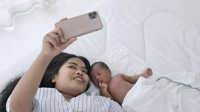 Mother and her newborn baby making a selfie or video call to the family on the bed white in a home. Mixed race black boy Ethnicity Thai-Nigeria. New generation Technology Communication