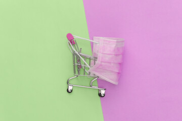 Mini supermarket trolley with medical mask on pink green pastel background. Top view