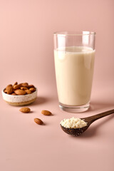 Vertical orientation almond milk in glass, flour in wooden brown spoon and nut kernels on pink powder background. Selective focus