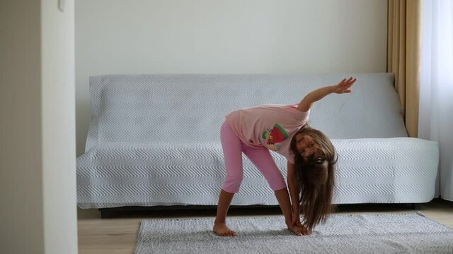 Funny little preschooler girl with long dark hair doing gymnastics exercises at home in living room near sofa and window, turning body to sides, stretching hips.
