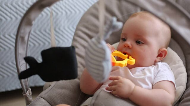 Cute little infant girl in white t shirt posing in bouncer looking at hanging toys and biting rattle, teething teeth, adorable newborn baby in rocking chair.