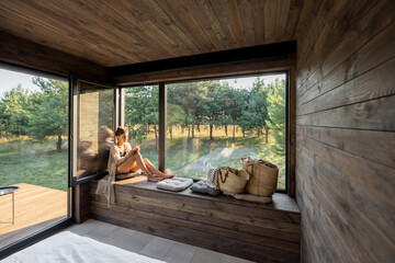 Young woman resting at beautiful country house or hotel, sitting with phone on the window sill enjoying beautiful view on pine forest. Concept of solitude and recreation on nature