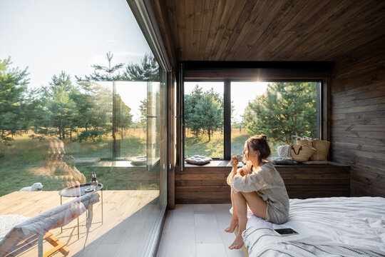 Woman in a country house or hotel with panoramic windows in pine forest sitting on the bed and enjoys beautiful view with light sun. Good morning and recreation on nature concept