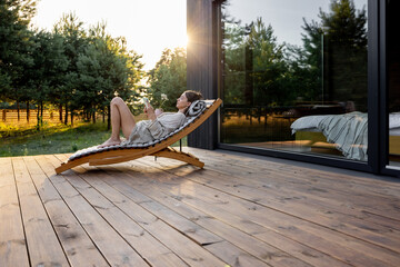 Young woman resting on sunbed and reading on a tablet on the wooden terrace near the modern house with panoramic windows near pine forest. Wellness and mindful health. Copy space