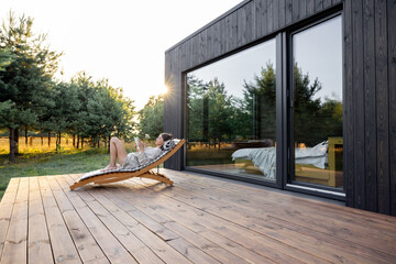 Fototapeta Young woman resting on sunbed and reading on a tablet on the wooden terrace near the modern house with panoramic windows near pine forest. Concept of solitude and recreation on nature obraz