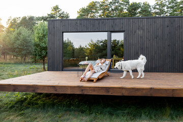 Woman resting on sunbed on the wooden terrace near the modern house with panoramic windows near pine forest with big white dog. Wellness and mindful health. Copy space