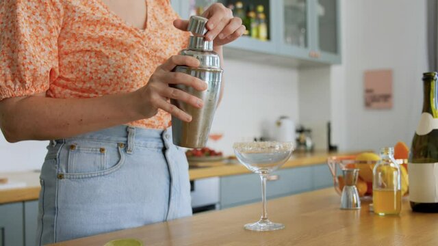 drinks and people concept - young woman with shaker pouring cocktail to glass at home kitchen