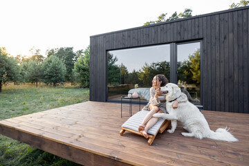 Woman enjoys the nature while sits on sunbed on wooden terrace near the modern house with panoramic windows near pine forest while hugs her pet. Concept of solitude and recreation on nature - 447487589