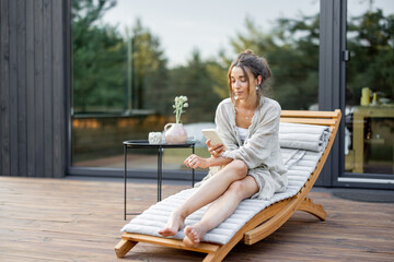 Young woman resting on sunbed and looks on the phone on terrace at modern house with panoramic windows near pine forest. Concept of solitude and recreation on nature. Wellness and mindful resort.