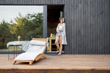 Foto op Canvas Young woman staying on terrace of wooden modern house with panoramic windows near pine forest. Concept of solitude and recreation on nature. Wellness and mindful resort. Beautiful place for vacation. © rh2010