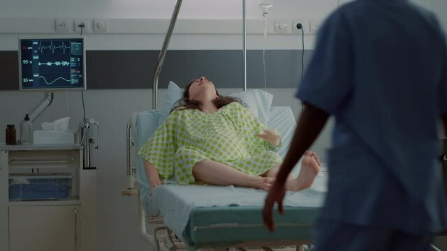 Young woman with pregnancy going into labor in hospital ward. Stressed husband bringing african american nurse for medical assistance and support against painful contractions at clinic