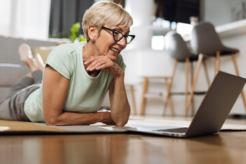Senior cheerful woman having fun while using computer on the floor in the living room