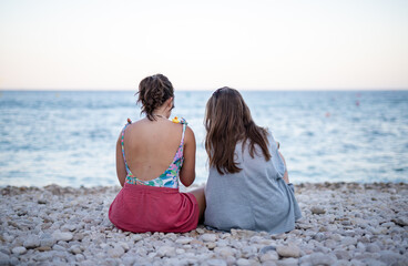 two young girl friends from the back staring at the sea sitting on rocks at a white beach