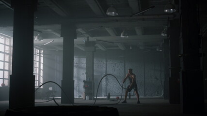 Guy battling ropes in gym. Athletic man doing crossfit workout in loft building