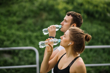 Young happy couple running in city park with plastic botle of water in hands, joint sports, cheerfulness, city sport healthy lifestyle, fitness together, runners, drinking water, thirst
