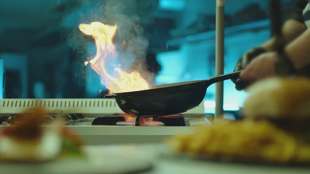 Professional chef cook flambe style . Prepares dish meat in a pan with open flames . Chef frying mixing food on gas hob in commercial kitchen . Man frying steak in flaming pan on hob in restaurant .  