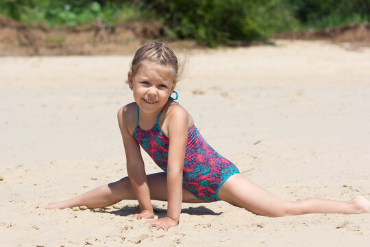 Caucasian little girl of five years old in swimsuit on sand beach making leg-split in summertime