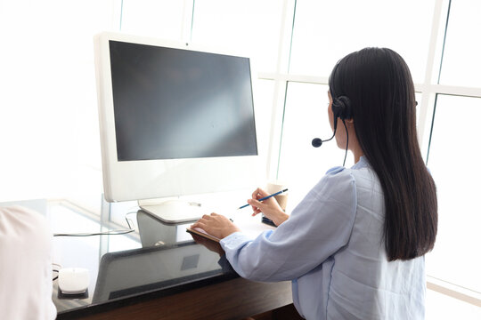 Asian Woman With Headphones Work At Call Center Service Desk Consultant From Behind, Call Center Agent With Headset Talk With Customer On Hotline, Operator With Hands-free Phone Has Conversation