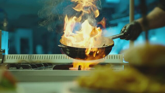 Professional chef cook flambe style . Prepares dish meat in a pan with open flames . Chef frying mixing food on gas hob in commercial kitchen . Man frying steak in flaming pan on hob in restaurant .  