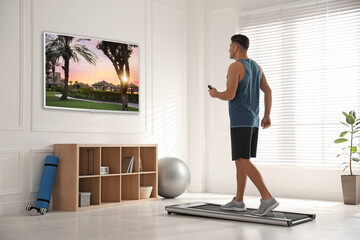 Fototapeta na wymiar Sporty man with remote control training on walking treadmill and watching TV at home