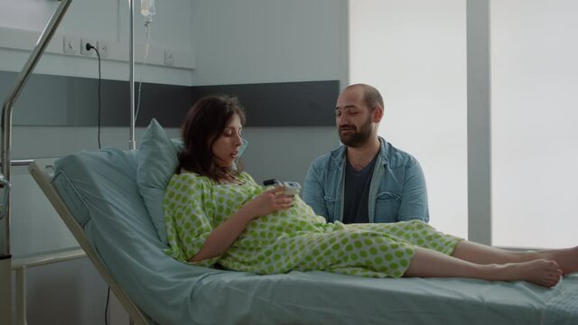 Young family talking about childbirth in hospital ward at maternity clinic. Caucasian pregnant woman laying in bed being happy about delivery. Husband sitting with wife for birth assistance