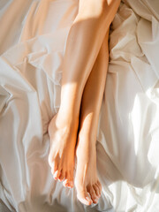 Morning at bed. Woman's feet on unmade bed. Sunbeams on white crumpled bed sheet. Light and shadow.