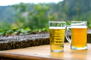 Two large mug of beer standing on a wooden table in the background a blurry river and valley.