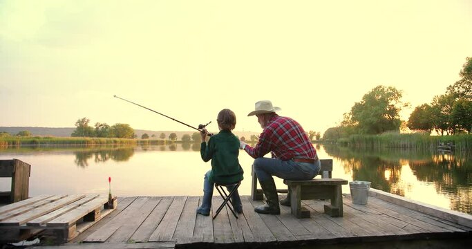 Rear view on the old grandfather and his small grandson fishing together and throwing rods in the lake while sitting on the wooden bridge or pier at the sunset
