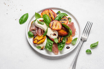 Grilled peach salad with mozzarella, prosciutto ham, basil, olive oil and balsamic. Fresh gourmet salad in plate on white stone background