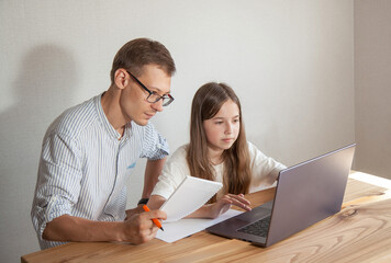 A teenage girl works on a laptop, a man is a teacher, a tutor or a dad with a notebook helps her