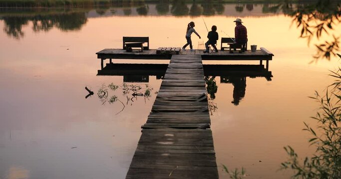 Back view of the happy family of three catching the fish at the pier during the sunset. Boy holding a fishing road and spending time with his family at the evening