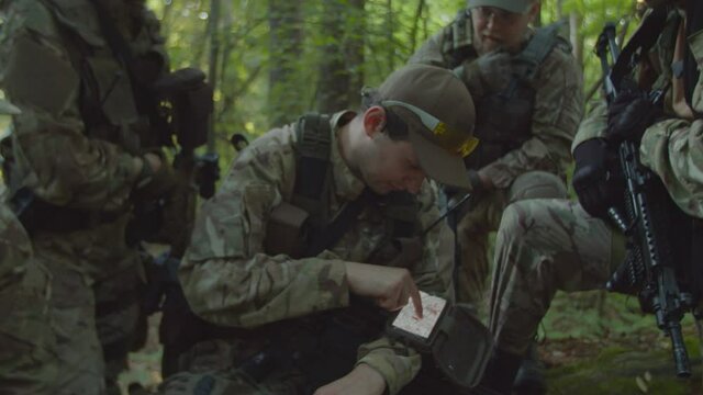 Detachment commander using army grade gadget, giving orders to fully armed squad of soldiers with rifles and machine gun in forest at daybreak. Military men and woman looking into digital map