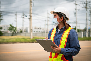 Woman who is a safety engineer with red safety vest and white hat on electric pole background.