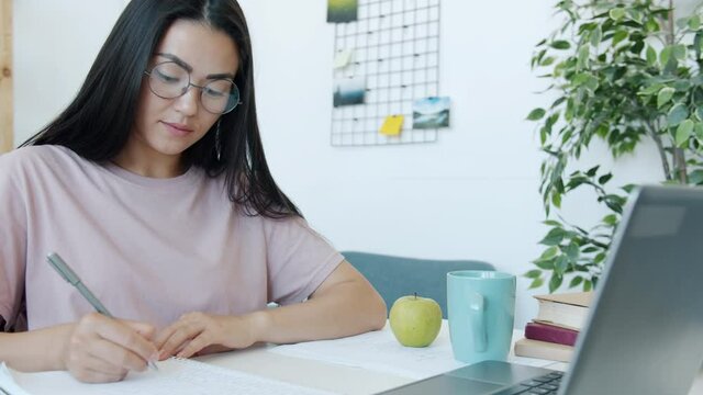 Slow motion of beautiful Asian woman student learning at home writing and looking at laptop screen sitting at teble in room. People and education concept.