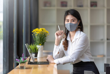 Asian businesswoman sitting at the office holding a stylus with a tablet at the office desk wear a mask to prevent germs. Look at the camera.