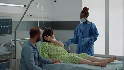 Caucasian couple expecting baby in maternity ward at hospital. Pregnant woman sitting in bed talking to african american nurse and young husband. Medical assistance for childbirth - Powered by Adobe