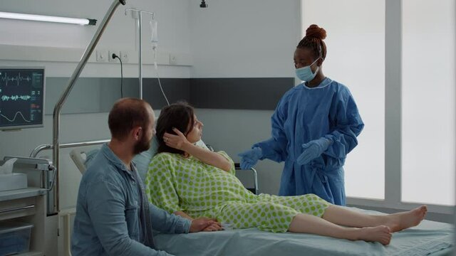 Caucasian couple expecting baby in maternity ward at hospital. Pregnant woman sitting in bed talking to african american nurse and young husband. Medical assistance for childbirth