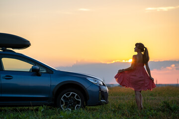 Dark silhouette of lonely woman relaxing near her car on grassy meadow enjoying view of colorful sunrise. Young female driver resting during road trip beside SUV vehicle.