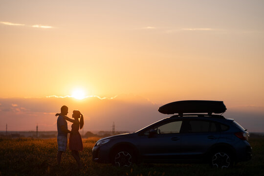 Happy couple relaxing beside their SUV car during honeymoon road trip at sunset. Young man and woman enjoying time together travelling by vehicle.