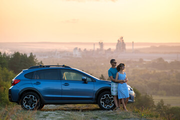 Happy couple standing near their car at sunset. Young man and woman enjoying time together travelling by vehicle.