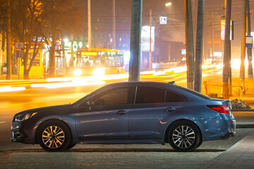 Obraz premium Close up of parked car on roadside at night with blurred view of traffic lights of moving vehicles on city street.