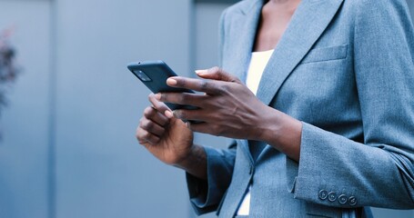 Selective focus, low angle view photo of classy adult woman holing cellular phone in hand, standing against blurred business center and checking something with smile